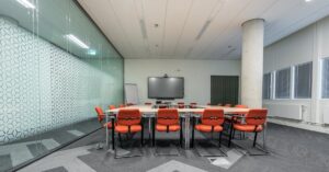 A modern conference room with a U-shaped table and orange cushioned chairs around it, facing a mounted screen on a white wall.