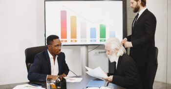 Three businessmen in a boardroom review documents as a colorful bar chart is shown on a large screen behind them.