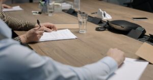Two professionals sign documents at a wooden conference table, with a glass of water and office equipment in view.