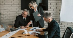 Three professionals in a modern office review documents at a conference table; a woman stands with a clipboard overseeing the work with two men at the table.