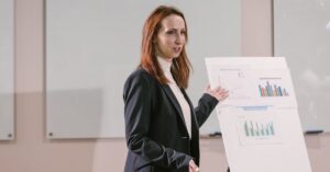 Woman in a dark blazer giving a business presentation with charts on a flip chart panel beside her