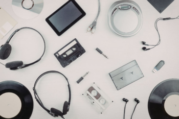 Image - Compliance Consultant London Flat lay of music tech on a white desk: headphones, vinyl records, a cassette, CDs, a tablet, cables, earphones, and a USB drive.
