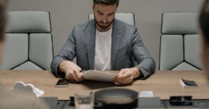 Man in a blazer studying documents at a conference table during a meeting.