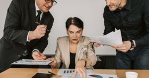 Three professionals review charts and documents spread on a desk during a meeting.