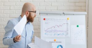 Side-profile of a bald man with a beard and glasses in a light blue shirt, studying printed charts pinned to a whiteboard with financial graphs.