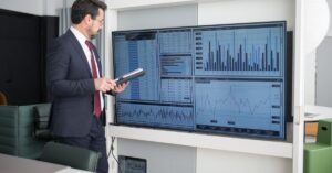 Businessman in a suit studies data dashboards on a large wall-mounted screen in a modern office with charts and graphs.
