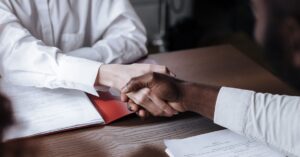 Two people shake hands over a desk with signed documents, signaling a business agreement.