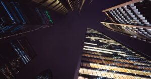 Nighttime cityscape with illuminated skyscrapers seen from the ground looking up at towering glass façades (worm's-eye view).