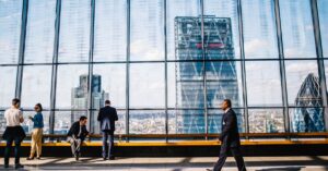 Professionals in business attire converse in a bright glass-walled atrium with a city skyline outside.