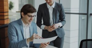 Two professionals in business attire examine a document together at a desk in a bright office environment.