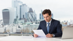 Businessman in a navy suit reviews documents at a desk with a laptop, urban skyline behind him.