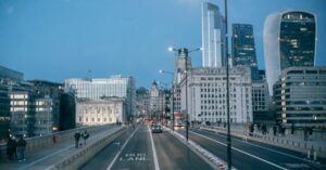 Evening city street on a bridge with a red double-decker bus and tall modern buildings in the background.