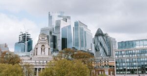 London skyline with a historic white building in the foreground and modern glass towers, including the Gherkin, behind trees under a cloudy sky.
