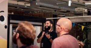 Photographer with a camera around his neck smiles while engaging with attendees at an indoor event, exposed ceiling and festive bunting in the background.