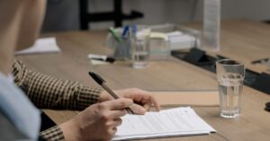 Person signing documents at a wooden desk with a pen, a glass of water nearby.