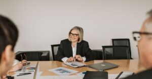 Businesswoman in a black blazer smiles while seated at a conference table with documents and a laptop in front of her.