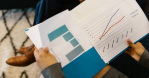 Person holds open report with a line graph showing two trends across days of the week; blue and orange lines rising over time.
