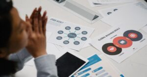Top-down view of a person with hands clasped at a white desk cluttered with business charts, a tablet, and printed sheets nearby a conference setup.